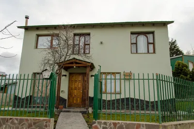 Casa de dos pisos con techo verde, puerta de madera y reja verde frente a un jardín con césped bajo un cielo nublado.