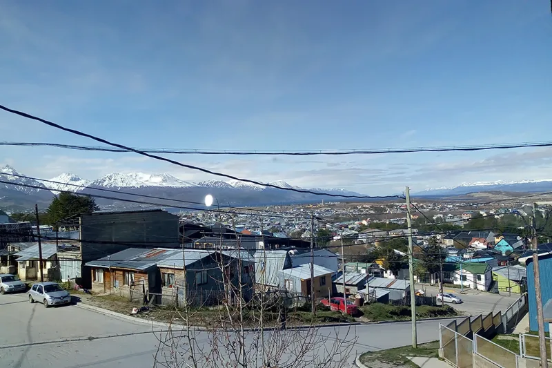 Vista urbana sobre casas de madera y techos de chapa con montañas nevadas al fondo bajo un cielo azul.
